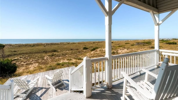A wooden deck with white railings overlooking a beach with dunes and the ocean.