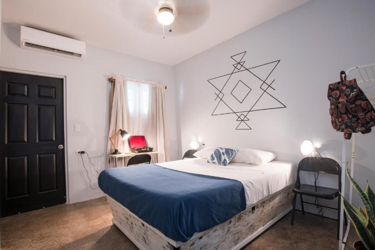 A bedroom with a bed, bedside lamps, a desk with a red lamp, a wall-mounted air conditioner, a door, and a window with curtains.