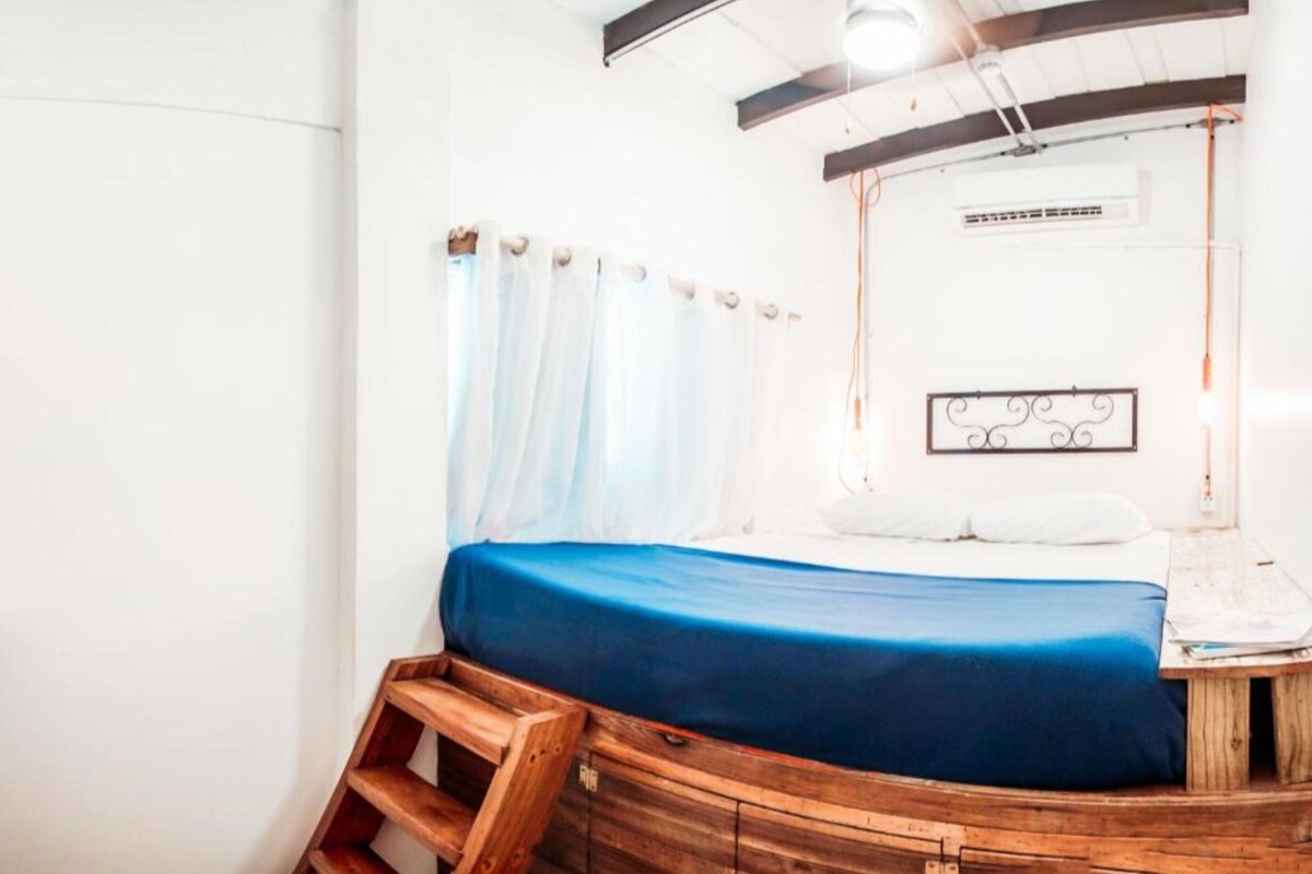 A room with a bed, a wooden bench, a white wall, a ceiling light, and a decorative metal piece.