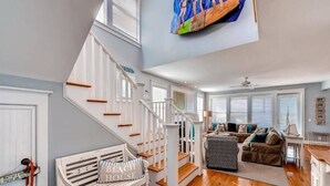 A spacious living area with a staircase leading to a second floor, a cozy seating area with a couch and armchairs, and a large window with blinds. - Footprints In The Sand 4 Bedroom Holiday Home By Bald Head Island (Bald Head Island)