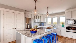 A kitchen with white cabinets, a marble island, blue stools, and a large window. - Hat Trick 4 Bedroom Holiday Home By Bald Head Island (Bald Head Island)