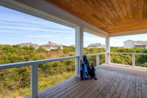 A covered outdoor deck with a wooden ceiling, white railings, and a view of houses and greenery in the background. - Hat Trick 4 Bedroom Holiday Home By Bald Head Island (Bald Head Island)