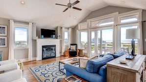 A spacious living room with a fireplace, a ceiling fan, and large windows offering a view of the outdoors. - Hat Trick 4 Bedroom Holiday Home By Bald Head Island (Bald Head Island)