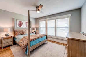 A bedroom with a bed, nightstands, a desk, a ceiling fan, and large windows with blinds.