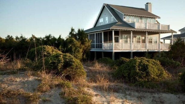 A two-story house with a balcony, surrounded by natural vegetation and sand.