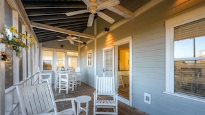 A covered porch with white Adirondack chairs and tables, a ceiling fan, and a view of the house in the background.