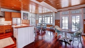 A spacious kitchen with wooden cabinetry and a large island, a dining area with a round table and chairs, and a bay window with multiple panes. - Island Belle 4 Bedroom Holiday Home By Bald Head Island (Bald Head Island)