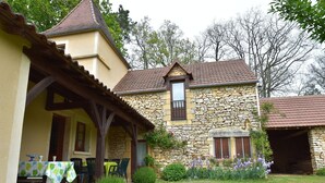 Ein Steingebäude mit Ziegeldach und einer Holzveranda. Vor der Veranda steht ein Tisch mit Stühlen, und ein kleiner Garten mit Blumen lädt zum Verweilen ein.