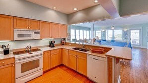 A modern kitchen with wooden cabinets, a microwave, stove, and dishwasher. It features a large window and a view of a balcony with outdoor seating. - Paradise 3 Bedroom Holiday Home By Bald Head Island (Bald Head Island)