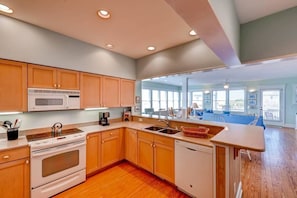 A modern kitchen with wooden cabinets, a microwave, stove, and dishwasher. It features a large window and a view of a balcony with outdoor seating.