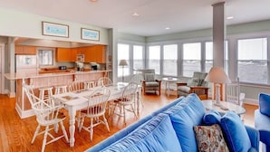 A spacious living area with a dining table and chairs, a blue sofa, and a view of the water through large windows. - Paradise 3 Bedroom Holiday Home By Bald Head Island (Bald Head Island)