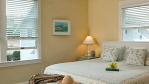 A bedroom with a bed, a nightstand, a lamp, and a window with blinds. - Purrfect Paws 2 Bedroom Holiday Home By Bald Head Island (Bald Head Island)