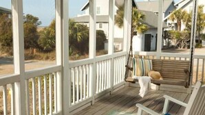 A wooden porch with a swing chair and a cushioned bench, overlooking a residential area with houses and palm trees. - Purrfect Paws 2 Bedroom Holiday Home By Bald Head Island (Bald Head Island)