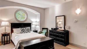 Une chambre à coucher comprenant un lit, des tables de chevet, une commode, une chaise, un ventilateur de plafond et une fenêtre ronde.