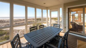 A covered outdoor dining area with a table and chairs, large windows offering a view of the beach, and a door leading to an adjacent room. - SeaWinds 4 Bedroom Holiday Home By Bald Head Island (Bald Head Island)