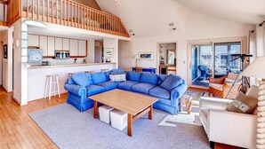 A spacious living area with a large blue sectional sofa, a wooden coffee table, and a white armchair. A kitchen with white cabinets and a wooden countertop is visible in the background. - SeaWinds 4 Bedroom Holiday Home By Bald Head Island (Bald Head Island)
