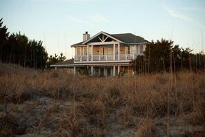 Uma casa de dois andares com varanda, cercada por gramíneas altas e árvores.