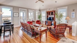 A spacious living room with hardwood flooring, a ceiling fan, a dining area with a table and chairs, a red sofa, a bookshelf, and a television. - Tiburon Marsh 4 Bedroom Holiday Home By Bald Head Island (Bald Head Island)