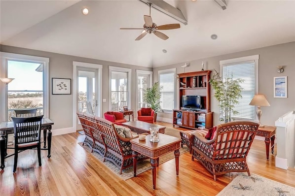 A spacious living room with hardwood flooring, a ceiling fan, a dining area with a table and chairs, a red sofa, a bookshelf, and a television.