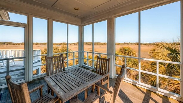 A covered outdoor seating area with a wooden table and chairs, large windows offering a view of the landscape. - Tiburon Marsh 4 Bedroom Holiday Home By Bald Head Island (Bald Head Island)