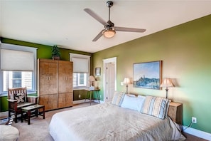 A bedroom with a bed, a chair, a wardrobe, a ceiling fan, and a painting on the wall.