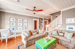 A living room with a beige sofa, a green coffee table, and a white armchair. There is a ceiling fan, framed pictures on the wall, and a staircase leading to another level.