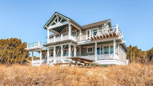 A large, two-story house with a prominent porch and multiple windows, surrounded by dry grass and trees. - Watch Hill 5 Bedroom Holiday Home By Bald Head Island (Bald Head Island)