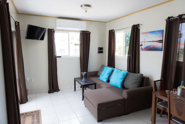 A living room with a brown sofa, a small table, a TV mounted on the wall, and a window with curtains.