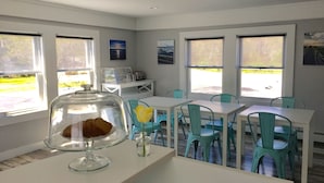 A dining area with white tables and teal chairs, a glass display case with a cake, and a view of a green landscape through large windows. - Kingfisher Lodging (Dennis)