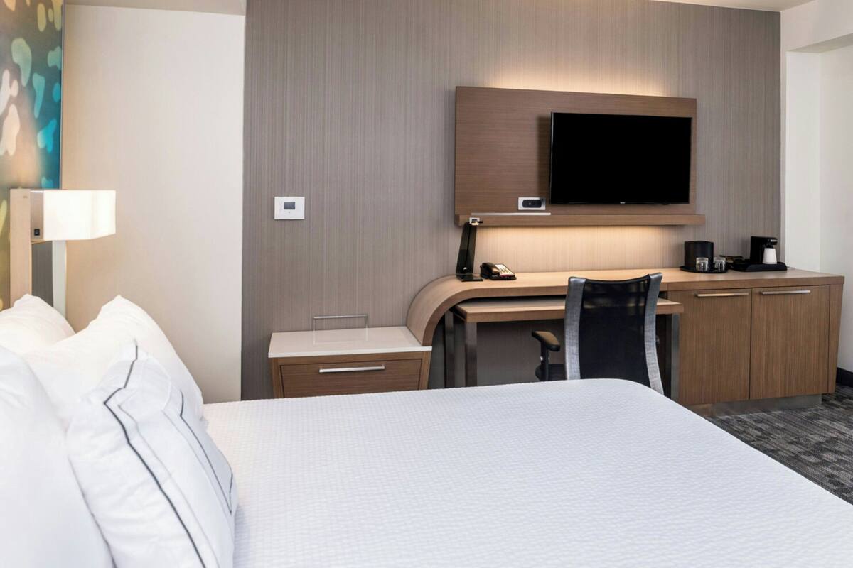 A hotel room with a large bed, a desk with a chair, a flat-screen TV mounted above the desk, and a coffee machine on the desk.