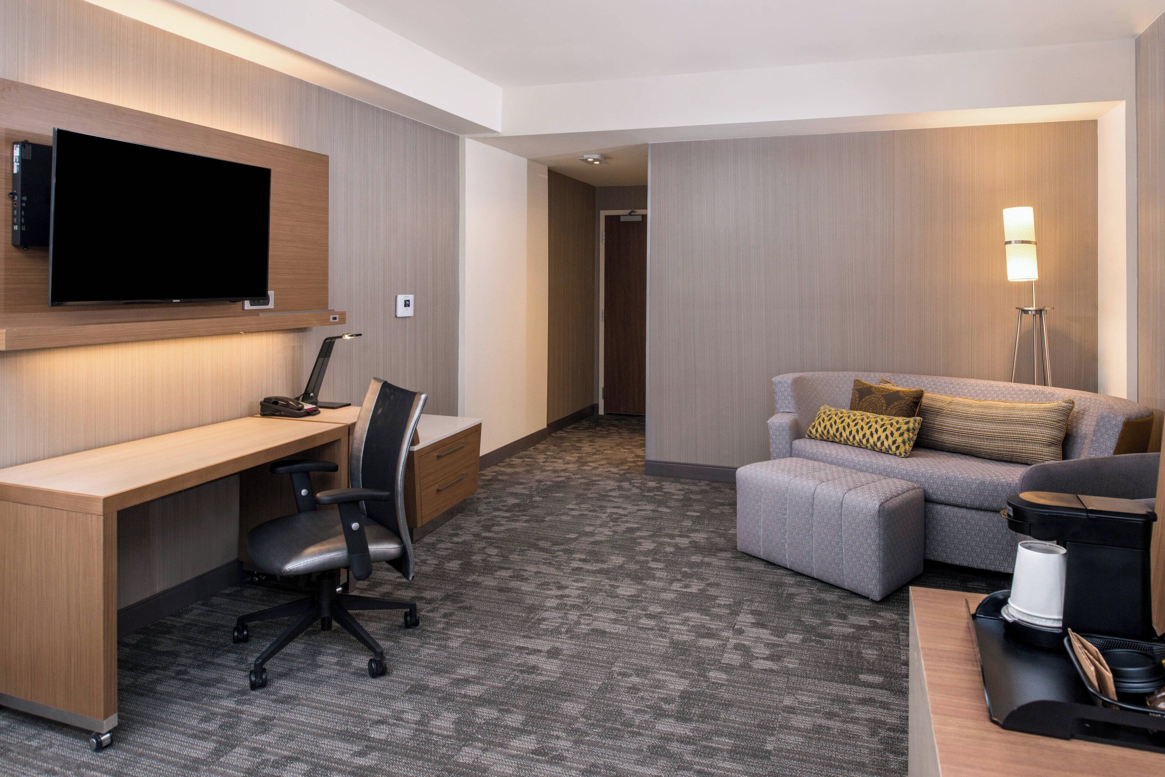 A hotel room with a flat-screen TV mounted on the wall, a desk with a chair, a sofa, and a coffee maker.