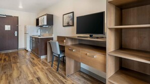 Une petite cuisine équipée d’un réfrigérateur, d’un four à micro-ondes et d’un évier, un bureau en bois avec une chaise, et un téléviseur posé sur un meuble.