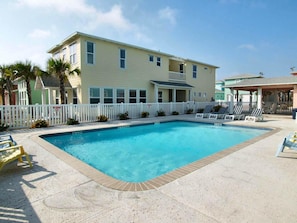 A pool area with a pool, lounge chairs, and a building with a white fence.