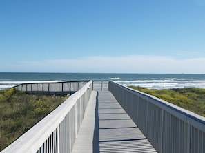 A wooden boardwalk leading to the ocean with a clear view of the sea and distant ships.
