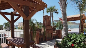 A wooden pergola with a picnic table, surrounded by palm trees and a stone fireplace. - Sun Castle PB221 (Port Aransas)
