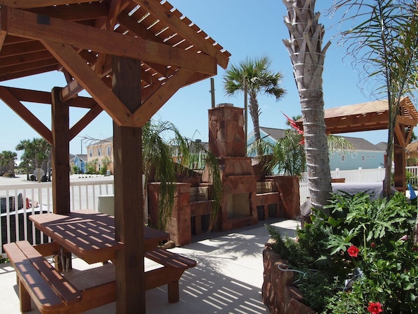 A wooden pergola with a picnic table, surrounded by palm trees and a stone fireplace.