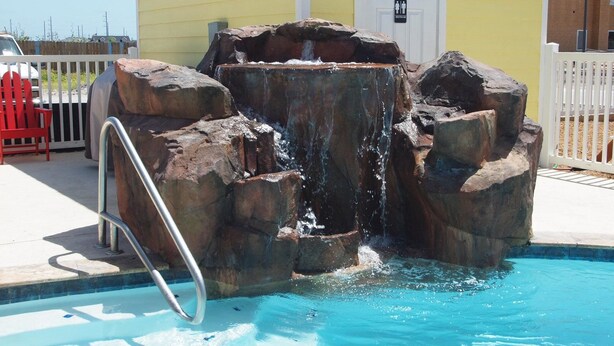 A pool with a rock waterfall feature, a red chair, and a sign indicating restrooms. - Sun Castle PB221 (Port Aransas)