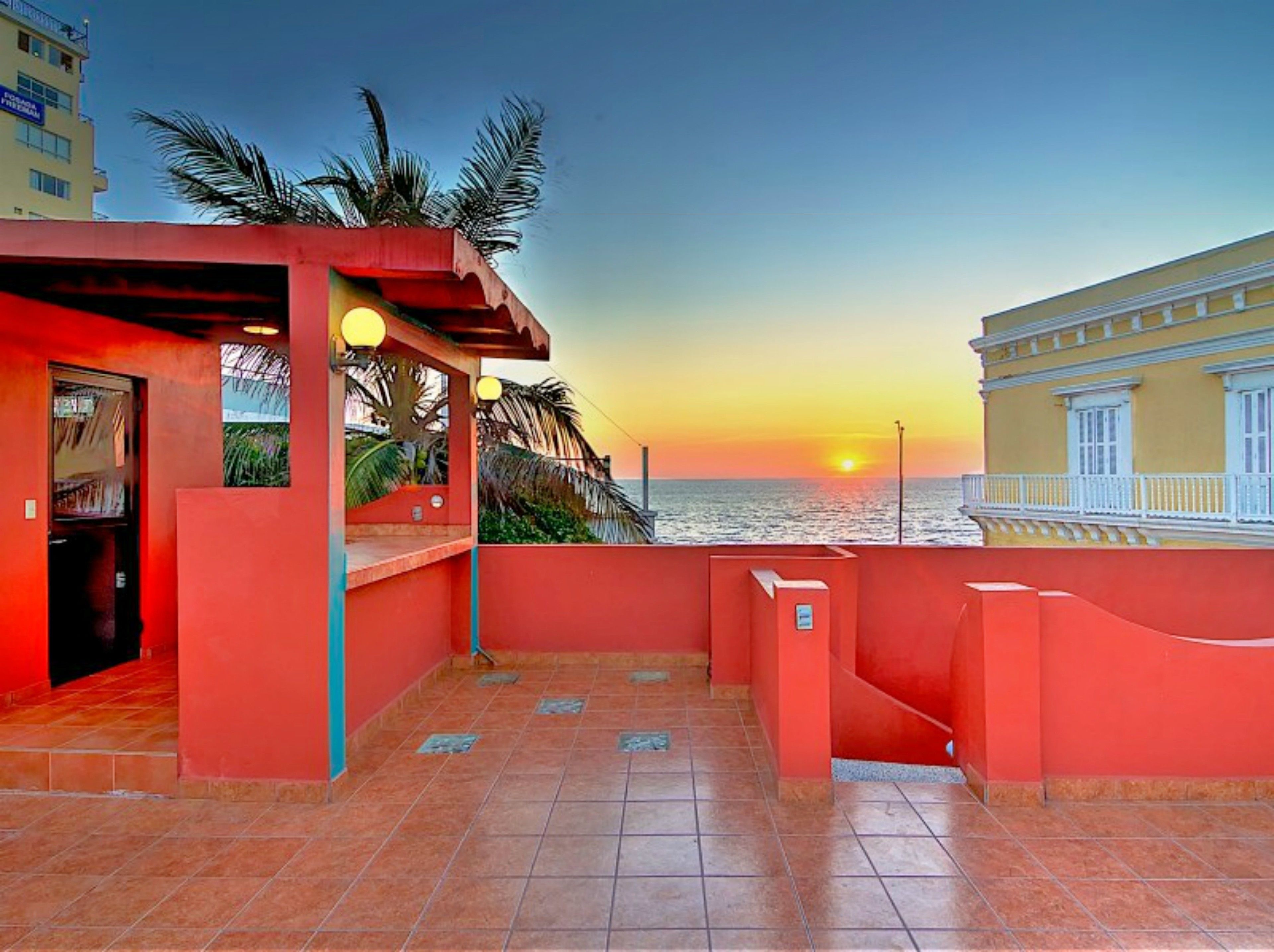 A red-roofed terrace with a view of the sunset over the sea, featuring a door and a small seating area with a railing.