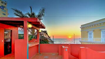 A red-roofed terrace with a view of the sunset over the sea, featuring a door and a small seating area with a railing.