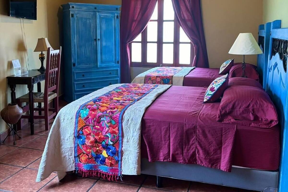 A bedroom with a bed, a ceiling fan, a window with curtains, a desk with a lamp, and a blue wardrobe.