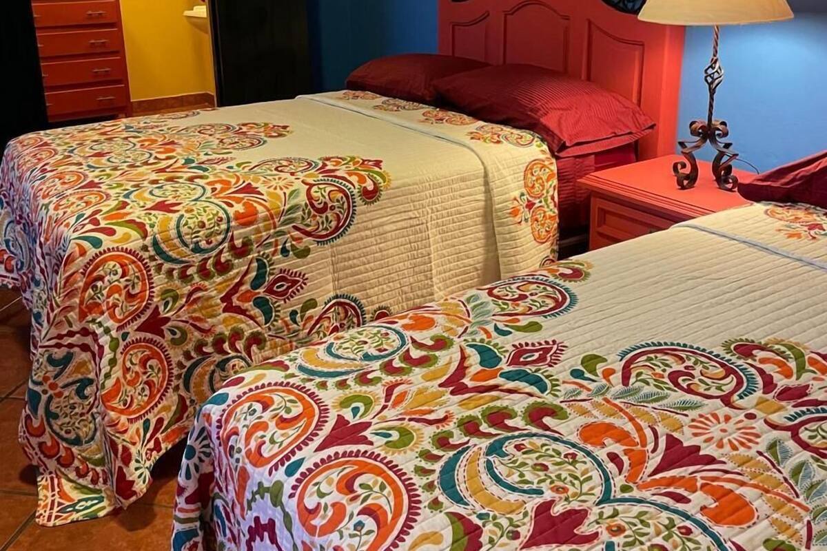 A room with two beds, each with a colorful patterned bedspread. There is a red nightstand with a lamp, and a wooden dresser with a mirror.
