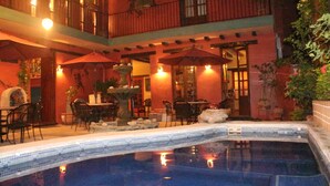 A pool area with a tiled edge, outdoor seating with umbrellas, and a building with a balcony.