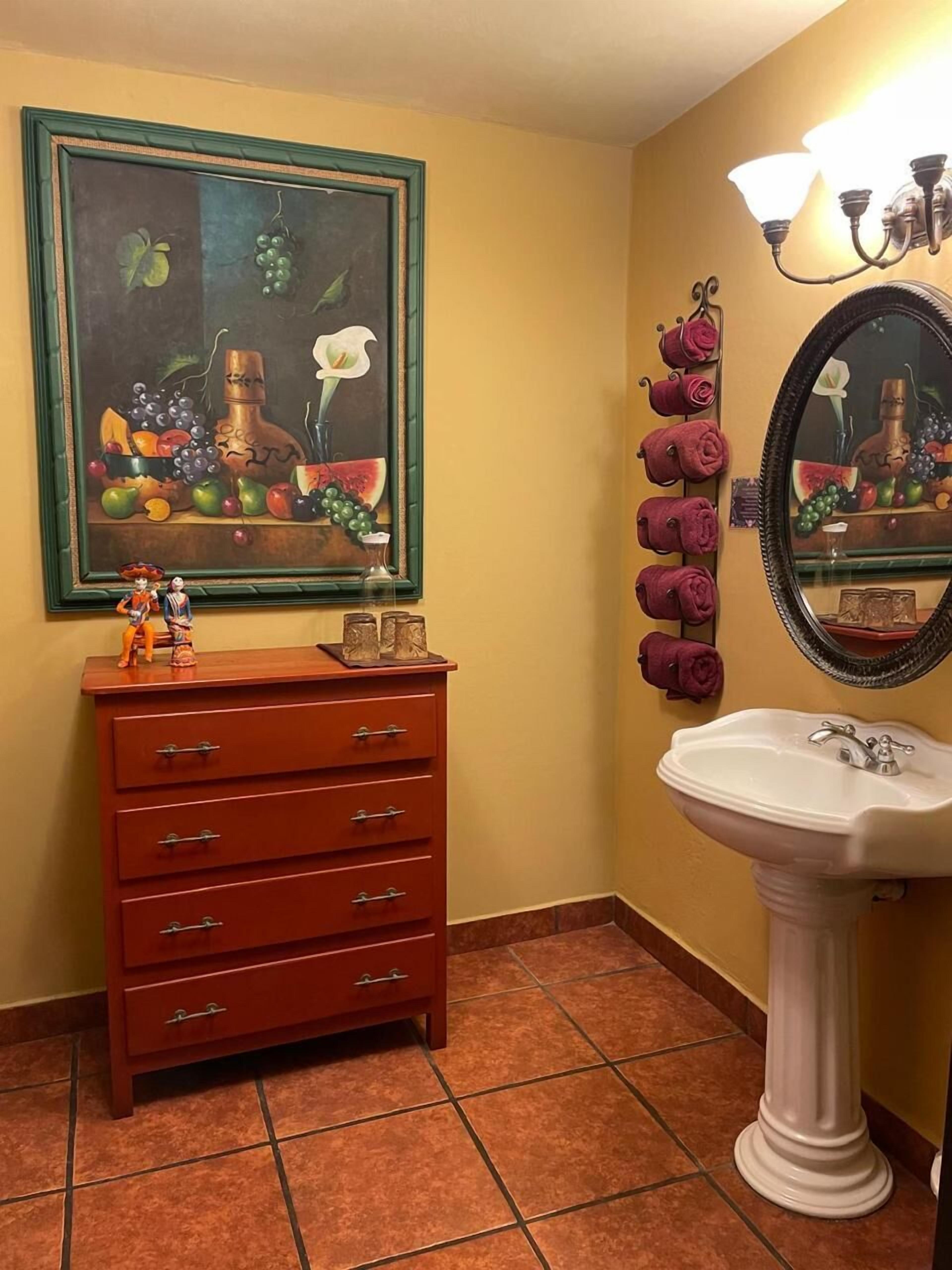 A bathroom with a wooden dresser, a white sink, a mirror with a light fixture, and a painting of fruits and flowers on the wall.