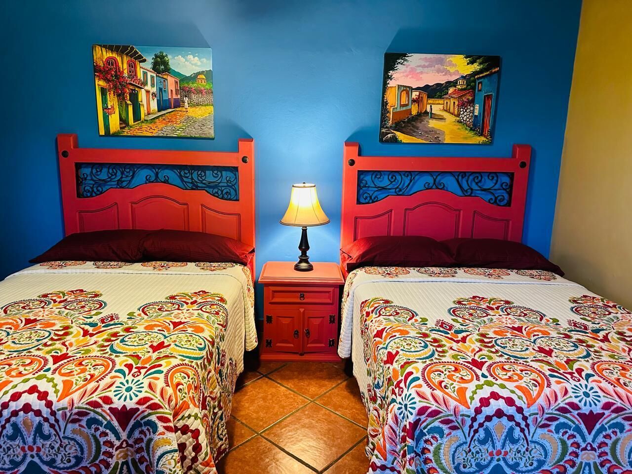 A room with two beds, each with a colorful patterned bedspread, a small nightstand with a lamp, and two wall-mounted paintings depicting colorful street scenes.
