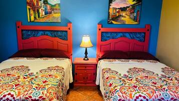 A room with two beds, each with a colorful patterned bedspread, a small nightstand with a lamp, and two wall-mounted paintings depicting colorful street scenes.