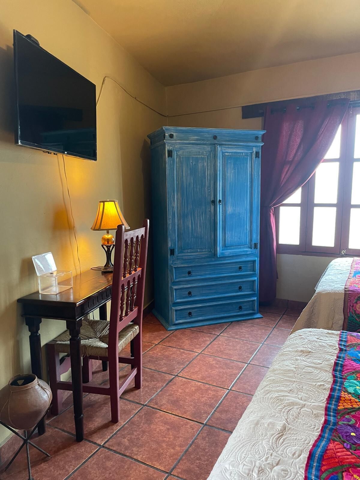 A room with a bed, a desk with a lamp, a blue cabinet, and a television mounted on the wall.