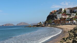 A coastal beach with buildings on a cliff, rocky shoreline, and calm sea. - Hotel Casa de Leyendas (Mazatlán)