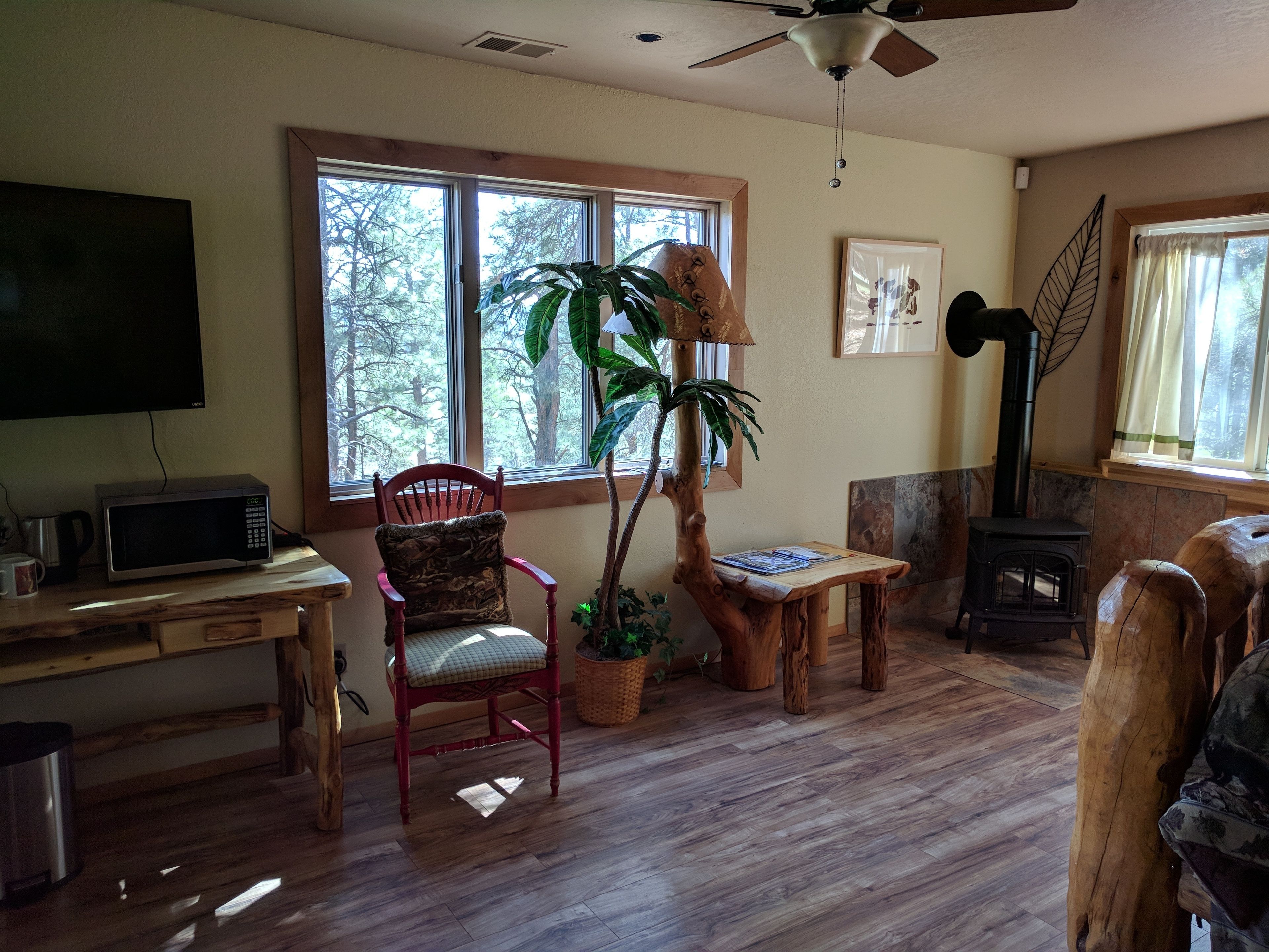 A cozy living room with a wooden floor, a red chair, a table with books, a fireplace, and a large window with a view of trees.