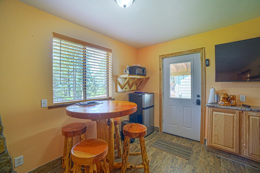 A compact kitchen with a wooden table and stools, a refrigerator, and a microwave on a small shelf.