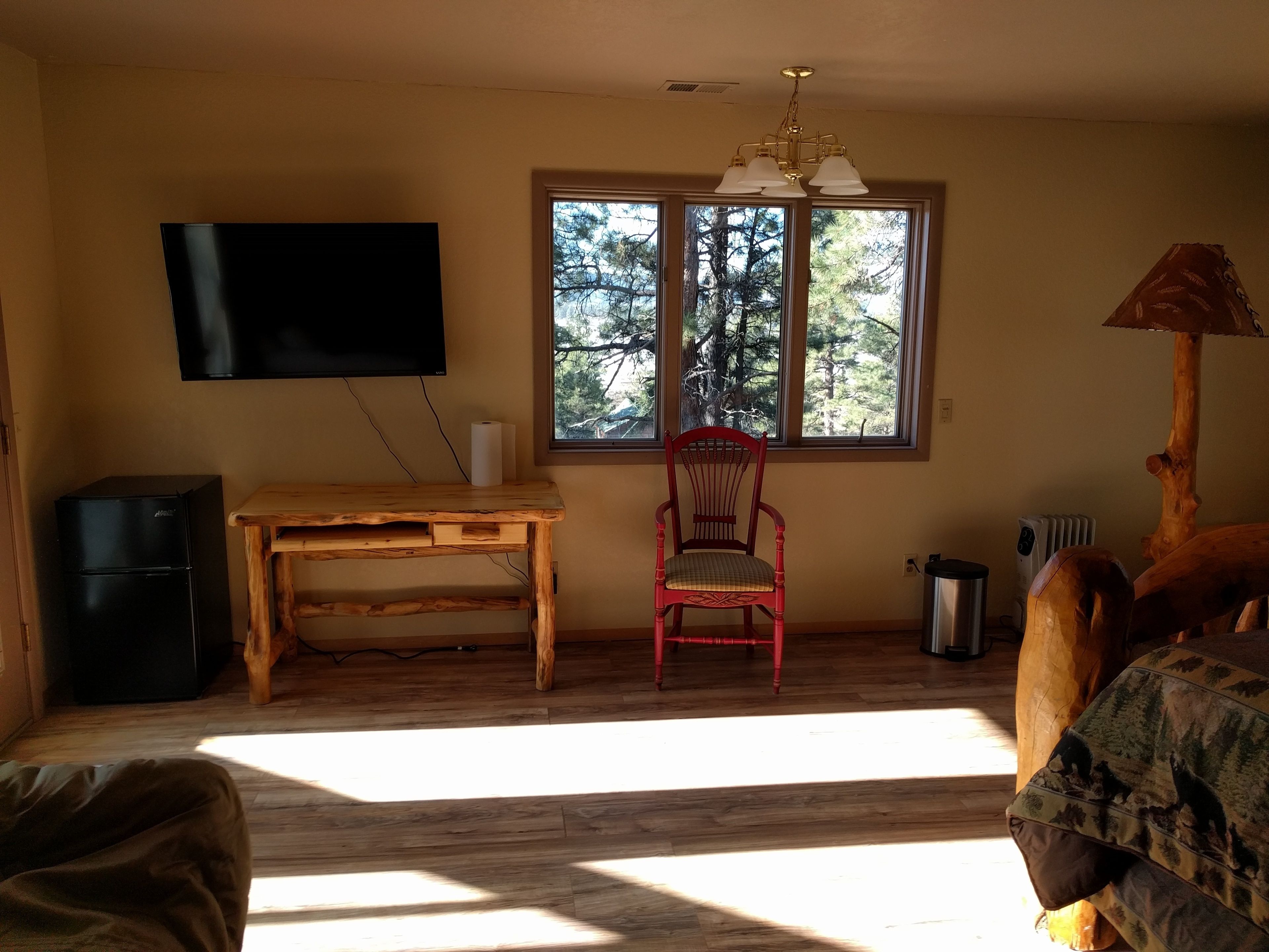 A room with a wooden desk, a red chair, a television mounted on the wall, and a window with a view of trees.